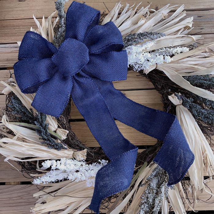 A rustic dried floral wreath with a large navy blue burlap bow, featuring beige dried grasses, white faux snow, and gray-green sprigs on a wooden plank background.