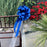 A large Santorini blue ribbon bow with long tails and a sheer white ribbon, tied to the back of a brown wooden chair in an outdoor garden setting with red flowers and greenery.