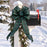A large green plaid holiday bow with a matching green garland and pinecones tied around a white mailbox post in a snowy outdoor setting.