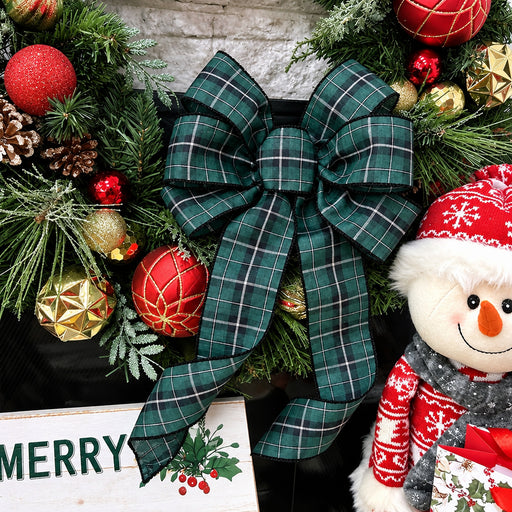 A large green plaid Christmas bow with black and white stripes, displayed on a black wreath decorated with red ornaments, pinecones, and evergreen branches, next to a snowman plush toy.