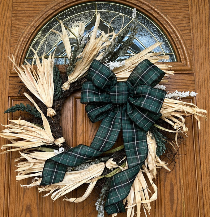 A rustic holiday wreath with dried palm fronds, evergreen branches, and white faux snow, decorated with a large dark green plaid bow, hanging on a brown wooden front door.