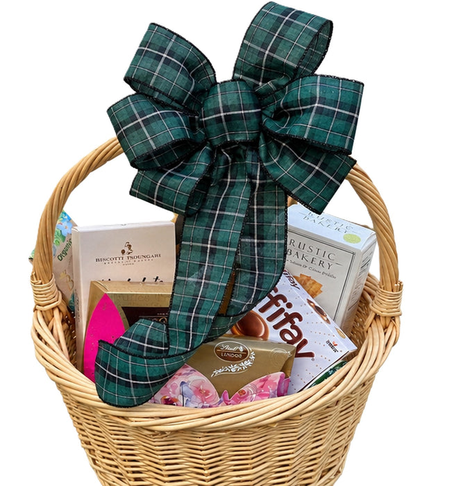 A wicker gift basket filled with assorted packaged snacks and treats, topped with a large green plaid bow. Visible items include a box of Brisket seasoning, a box of Rustic Bakery cookies, a bag of Ritz crackers, and other candy and snack packages, all photographed on a plain white background.