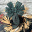 A large green plaid bow with white windowpane lines, attached to a rustic holiday wreath made of dried grasses, evergreen branches, and white floral accents, set against a light gray brick wall.