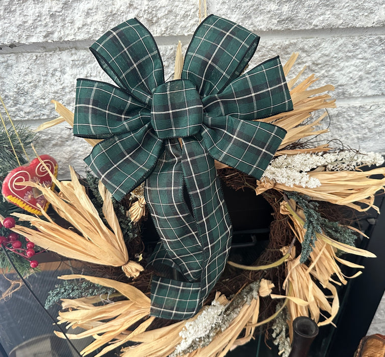 A large green plaid bow with white windowpane lines, attached to a rustic holiday wreath made of dried grasses, evergreen branches, and white floral accents, set against a light gray brick wall.