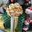 A large decorative gift bow in gold and cream is tied around a green plaid Christmas gift box, with snowman decorations and a holiday tree in the background.