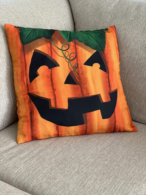 A square decorative throw pillow featuring a carved jack-o'-lantern face design in orange, black, and green, resting on a textured gray fabric sofa.